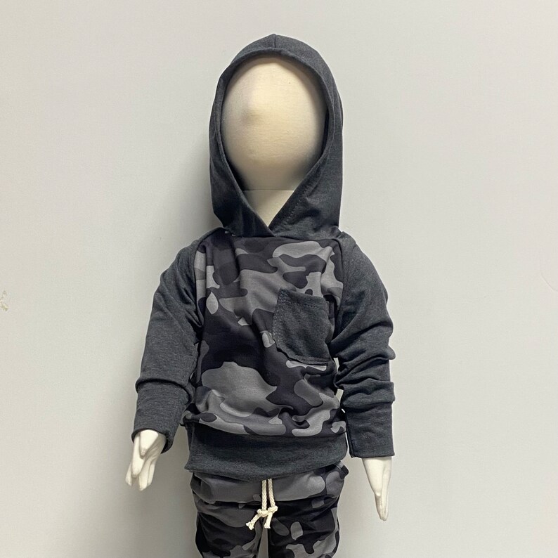 May include: A gray hooded sweatshirt with a camouflage pattern on the front. The sweatshirt has a pocket on the front and a drawstring hood.