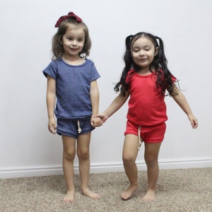 May include: Two young girls wearing matching short-sleeved shirts and shorts. The girl on the left is wearing a blue shirt and shorts, while the girl on the right is wearing a red shirt and shorts. Both girls are smiling and holding hands.