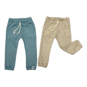 May include: Two pairs of children's sweatpants. One pair is teal, the other is tan. Both have an elastic waistband with a drawstring, front pockets, and a small logo near the ankle. The pants are made of a soft, comfortable material.