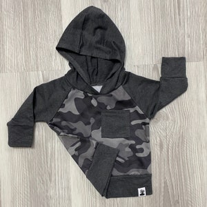 May include: A gray hooded sweatshirt with a camouflage pattern on the front and a small pocket on the left side. The sweatshirt has long sleeves and a drawstring hood.