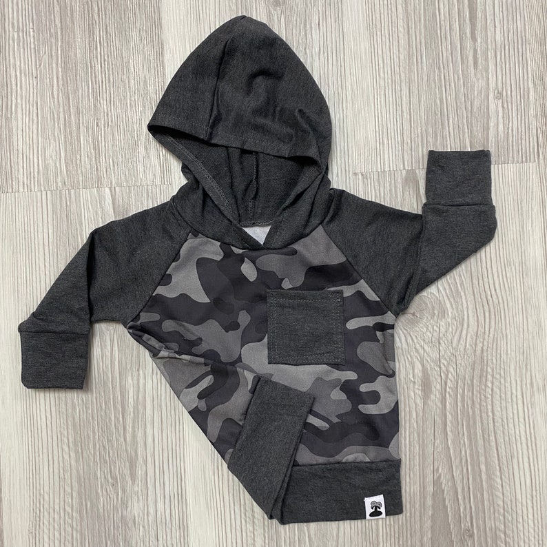 May include: A gray hooded sweatshirt with a camouflage pattern on the front and a small pocket on the left side. The sweatshirt has long sleeves and a drawstring hood.