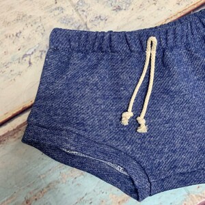 May include: Blue denim shorts with a drawstring waist. The drawstring is made of a cream-colored fabric.