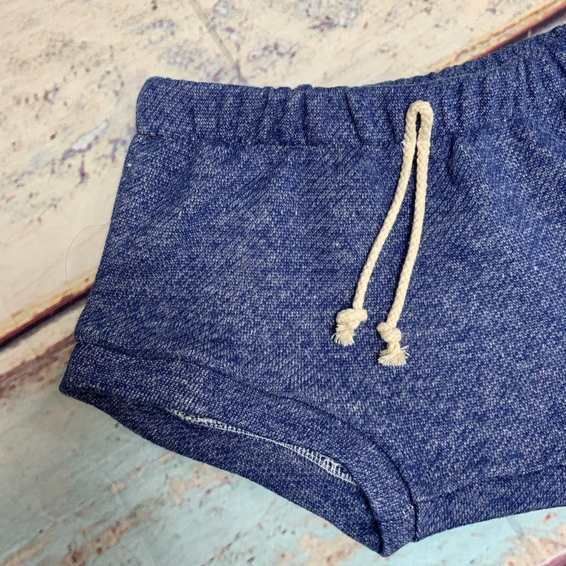 May include: Blue denim shorts with a drawstring waist. The drawstring is made of a cream-colored fabric.