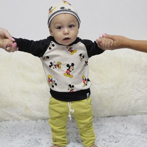 May include: A baby in a beige and black Mickey Mouse print sweatshirt, matching beanie, and yellow sweatpants. The outfit has black sleeves and a black bottom hem. The sweatpants have a drawstring. The baby is standing on a white fluffy rug.
