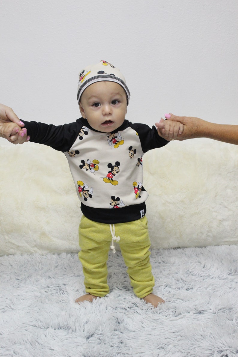 May include: A baby in a beige and black Mickey Mouse print sweatshirt, matching beanie, and yellow sweatpants. The outfit has black sleeves and a black bottom hem. The sweatpants have a drawstring. The baby is standing on a white fluffy rug.