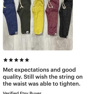 May include: Five pairs of sweatpants in navy blue, olive green, yellow, burgundy, and charcoal grey. Each pair has a drawstring waist and a small white tag. The pants are displayed on a light wood surface.