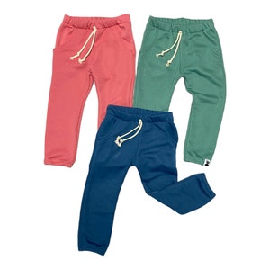May include: Three pairs of children's sweatpants in coral, teal, and green. Each pair has a drawstring waist and front pockets. The pants are made of a soft material and are suitable for casual wear.