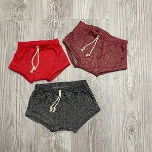 May include: Three pairs of toddler shorts in different colors. The shorts are made of a soft, stretchy fabric and have a drawstring waistband. The colors are red, burgundy, and gray.