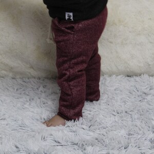 May include: A child in a black top and burgundy trousers made of textured knit fabric. A small white tag with a tree logo is visible on the top. The child is standing on a fluffy white rug. The trousers are a deep red colour.
