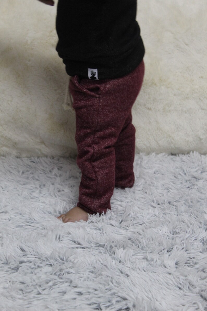 May include: A child in a black top and burgundy trousers made of textured knit fabric. A small white tag with a tree logo is visible on the top. The child is standing on a fluffy white rug. The trousers are a deep red colour.