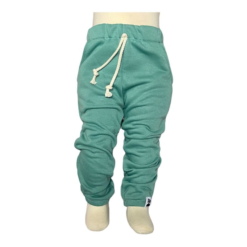 May include: Teal-colored baby sweatpants with a drawstring waist and gathered ankles. The pants have a relaxed fit and a small white tag on the lower leg. The drawstring is a cream-colored rope.