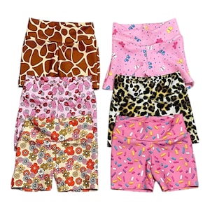 May include: A collection of six pairs of athletic shorts with various patterns. The patterns include giraffe print, pink with butterflies, pink with ice cream, leopard print, floral, and pink with sprinkles. The shorts are made of a textured fabric.