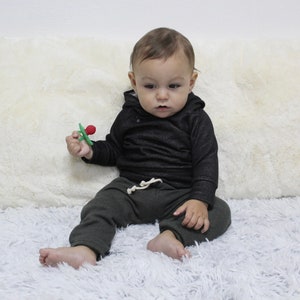May include: A baby in a black hooded top and dark green sweatpants with a white drawstring. The baby holds a green and red teething toy. The baby is sitting on a fluffy white surface.