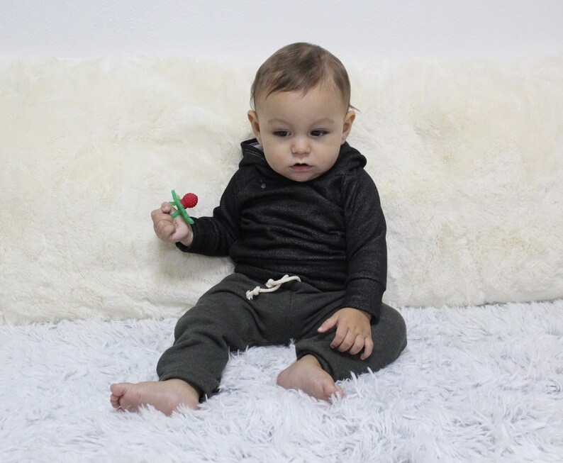 May include: A baby in a black hooded top and dark green sweatpants with a white drawstring. The baby holds a green and red teething toy. The baby is sitting on a fluffy white surface.