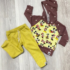 May include: A children's outfit comprising mustard yellow sweatpants and a maroon hooded sweatshirt. The sweatshirt has white polka dots on the sleeves and hood, and a yellow panel with Mickey Mouse and Minnie Mouse graphics.