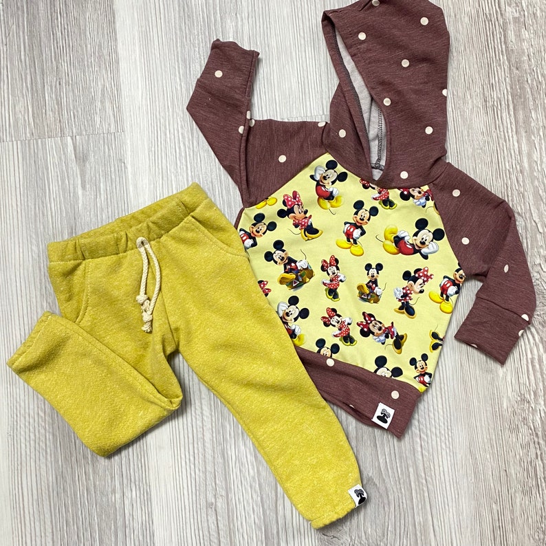 May include: A children's outfit comprising mustard yellow sweatpants and a maroon hooded sweatshirt. The sweatshirt has white polka dots on the sleeves and hood, and a yellow panel with Mickey Mouse and Minnie Mouse graphics.
