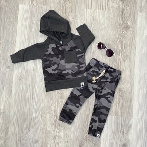 May include: A gray and black camouflage hoodie and matching sweatpants for a child. The hoodie has a drawstring hood and a small pocket on the front. The sweatpants have a drawstring waistband and a small logo on the leg.