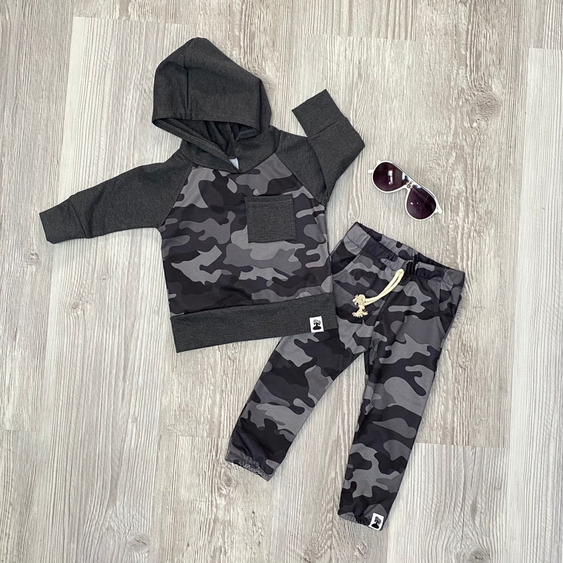 May include: A gray and black camouflage hoodie and matching sweatpants for a child. The hoodie has a drawstring hood and a small pocket on the front. The sweatpants have a drawstring waistband and a small logo on the leg.
