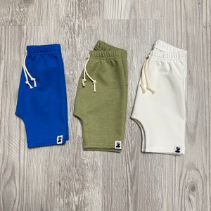 May include: Three pairs of shorts in blue, olive green, and white. Each pair has a drawstring waist and a small logo patch. The shorts are made of a soft fabric and are suitable for casual wear.