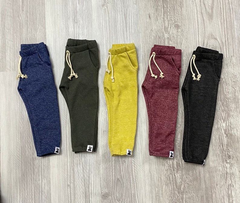 May include: Five pairs of children's sweatpants in navy blue, olive green, mustard yellow, burgundy, and charcoal grey. Each pair has a drawstring, pockets, and a small white label. The trousers are displayed on a light wood-grain surface.