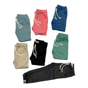 May include: A collection of folded sweatpants in various colors, including teal, coral, navy, tan, black, and green. Each pair features a drawstring waist and a front pocket. The pants are made of a soft material.