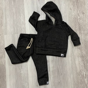May include: A black hooded sweatshirt and matching sweatpants set. The hoodie has a drawstring and a kangaroo pocket. The sweatpants have a drawstring and pockets. Both pieces have a small white tag with a graphic on the lower left.