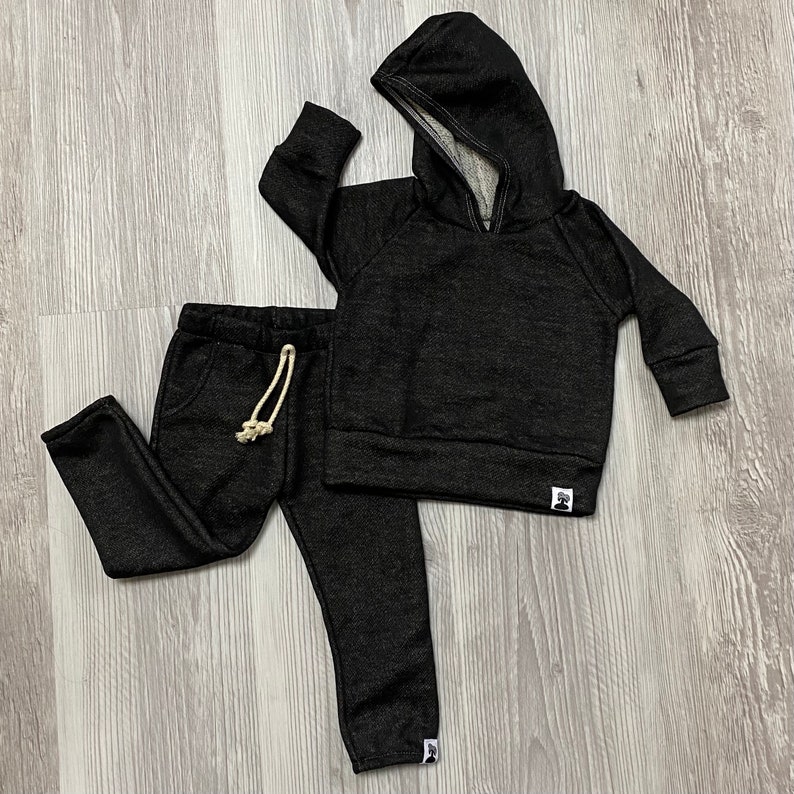 May include: A black hooded sweatshirt and matching sweatpants set. The hoodie has a drawstring and a kangaroo pocket. The sweatpants have a drawstring and pockets. Both pieces have a small white tag with a graphic on the lower left.