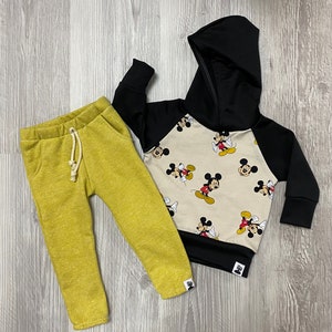 May include: A children's two-piece outfit. The outfit includes yellow-green sweatpants and a black and beige Mickey Mouse hooded sweatshirt. The sweatshirt has black sleeves and hood, with a beige body covered in Mickey Mouse graphics.