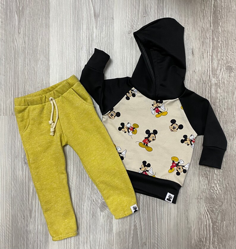 May include: A children's two-piece outfit. The outfit includes yellow-green sweatpants and a black and beige Mickey Mouse hooded sweatshirt. The sweatshirt has black sleeves and hood, with a beige body covered in Mickey Mouse graphics.