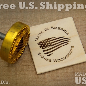 May include: A gold-colored metal branding iron with a wooden handle. The branding iron has a design of an American flag with stars and stripes. The text "MADE IN AMERICA" and "SPARKS WOODWORKS" is around the flag design. The text "MADE IN USA" is on a separate piece of wood.