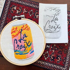 May include: Embroidered artwork of a La Croix can in a wooden hoop. The can design features yellow, pink, blue, and orange colors. The words "La Croix" are embroidered in blue. A printed image of the can is next to the embroidery.