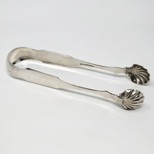 May include: Silver sugar tongs with shell-shaped handles. The tongs are in a closed position.
