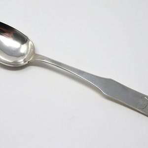 May include: A silver spoon with a rounded bowl and a long, slender handle. The handle has a simple, rounded design and is engraved with the initials "J.C."