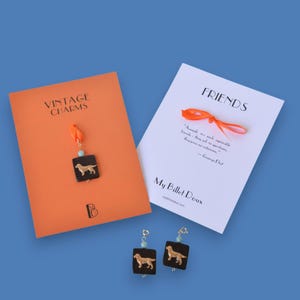 May include: Earrings with a golden retriever design on a black square, hanging from a gold-colored hook with a blue bead. The earrings are displayed with a card that says "VINTAGE CHARMS" and another card that says "FRIENDS".