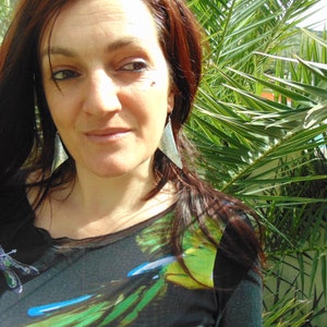 May include: A woman with long, dark brown hair wearing a black and green patterned shirt with a dragonfly design. She is wearing large silver triangle earrings.