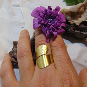 May include: A gold hammered ring with a wide band. The ring is on a finger and the hand is resting on a piece of driftwood.