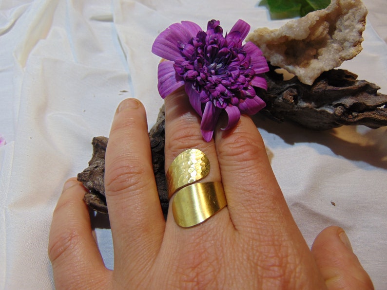 May include: A gold hammered ring with a wide band. The ring is on a finger and the hand is resting on a piece of driftwood.