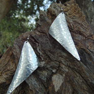 May include: A pair of silver triangle earrings with a hammered texture. The earrings are hanging from a wooden surface.