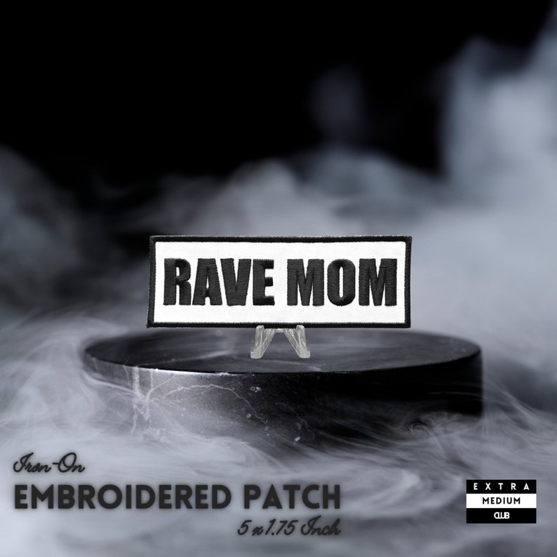 May include: An iron-on embroidered patch with the words "RAVE MOM" in black block letters on a white background. The patch measures 5 x 1.75 inches. The patch is displayed on a small clear stand against a smoky background.
