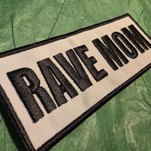 May include: Embroidered patch with the words "RAVE MOM" in bold, black letters on a white background. The patch has a black border and is set against a green, textured surface. The patch is rectangular.