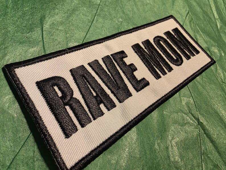 May include: Embroidered patch with the words "RAVE MOM" in bold, black letters on a white background. The patch has a black border and is set against a green, textured surface. The patch is rectangular.