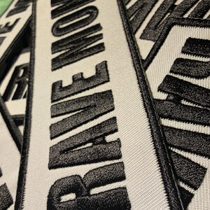 May include: Close-up of several rectangular white patches with the word "RAVE NOW" embroidered in bold black letters. The patches have a black border and are made of a textured fabric.