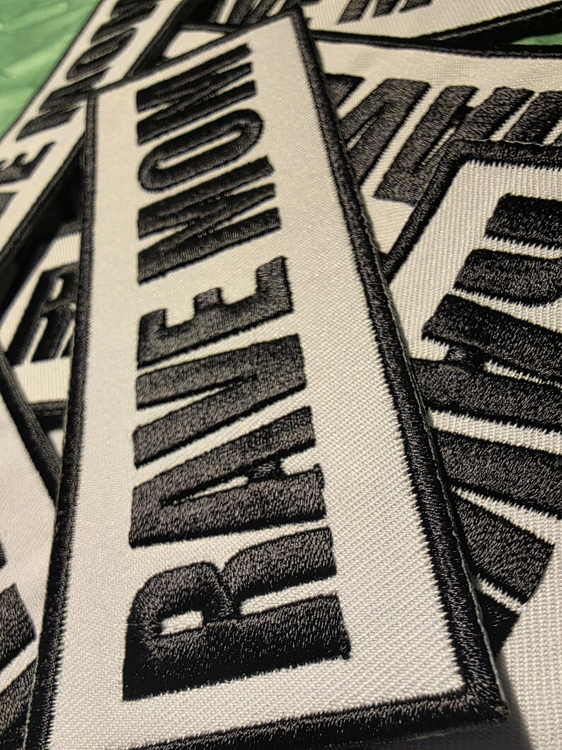 May include: Close-up of several rectangular white patches with the word "RAVE NOW" embroidered in bold black letters. The patches have a black border and are made of a textured fabric.