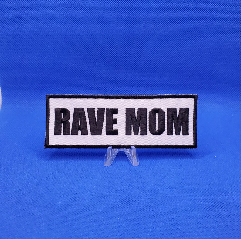 May include: A rectangular patch with the words "RAVE MOM" embroidered in black on a white background. The patch has a black border and is displayed on a clear plastic stand against a blue backdrop.