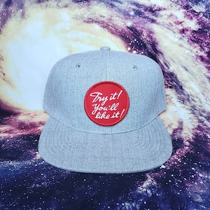 Puede incluir: Gorra de béisbol gris con visera plana y parche circular rojo. El parche presenta el texto blanco "Try it! You'll like it!". La gorra tiene un botón en la parte superior y está sobre un fondo con tema de galaxia.