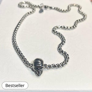 Puede incluir: Un collar de cadena de plata con un dije de calavera. La cadena está hecha de pequeños eslabones redondos. El dije de calavera es detallado y tiene un aspecto realista.
