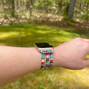 May include: A silver Apple Watch with a colorful beaded band. The band is made of multiple rows of small, colorful beads in shades of blue, red, white, and green. The watch is worn on a person's wrist.