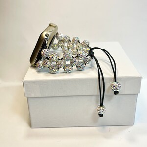 May include: A silver Apple Watch with a white crystal beaded band and black cord with crystal beads at the ends. The watch is on a white box.