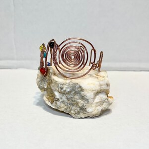 May include: A copper wire sculpture of a spiral with colorful beads. The sculpture is sitting on a white rock.