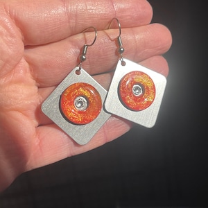 May include: A pair of silver square earrings with a red and orange glitter circle in the center. Each circle has a clear rhinestone in the center.